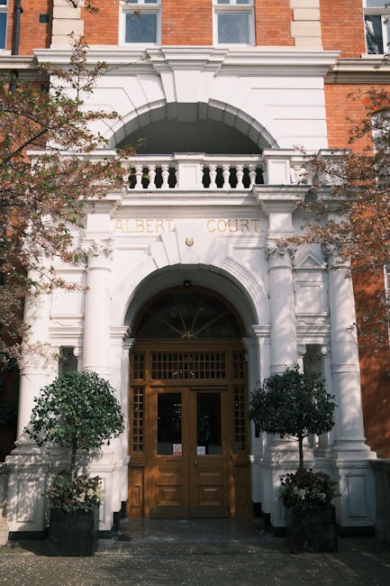 The image depicts the entrance to a building labeled 'Albert Court,' featuring a white stone facade with classical architectural details. The entrance has a large, rounded archway with a wooden double