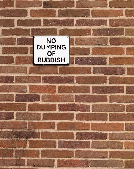 A red, metal waste container with a weathered, chipped surface is positioned against a dark, textured wall and a light grey, rough concrete wall in an urban outdoor setting. Next to the container, mul