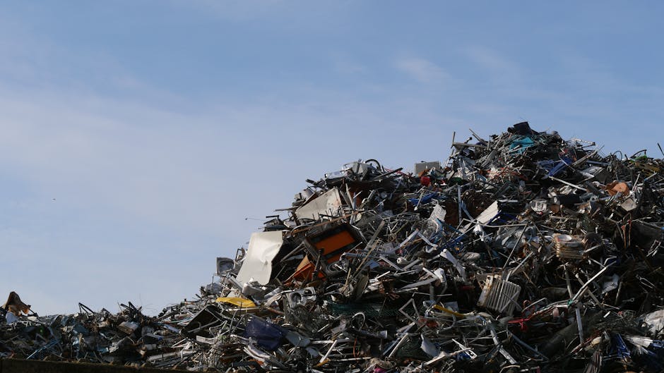 A large collection of mixed household waste and discarded items scattered on the ground amidst dry, brown grass and leafless branches. The debris includes cardboard boxes, plastic containers, a microw