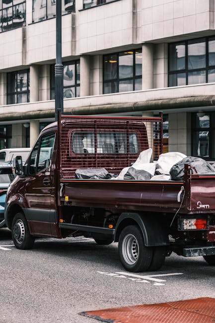 A burgundy flatbed utility truck parked on an urban street, loaded with various waste materials including large black and gray bags, some covered with plastic sheeting, and loose debris in the truck b