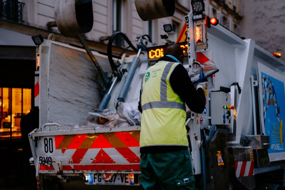 A burgundy flatbed utility truck parked on an urban street, loaded with various waste materials including large black and gray bags, some covered with plastic sheeting, and loose debris in the truck b